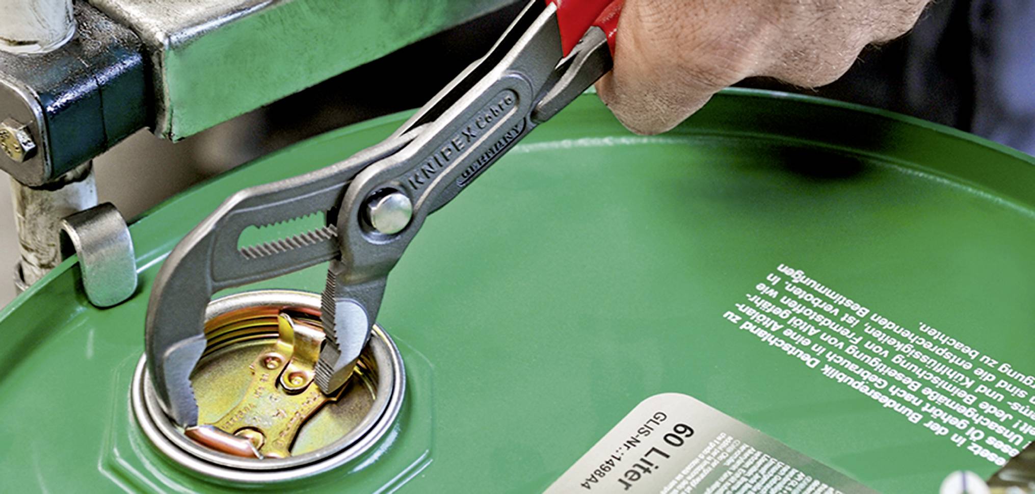 Hand benutzt Zange, um den Verschluss eines grünen 60-Liter-Fasses zu öffnen. Fokussiert auf Werkzeug und Mechanismus des Fasses.