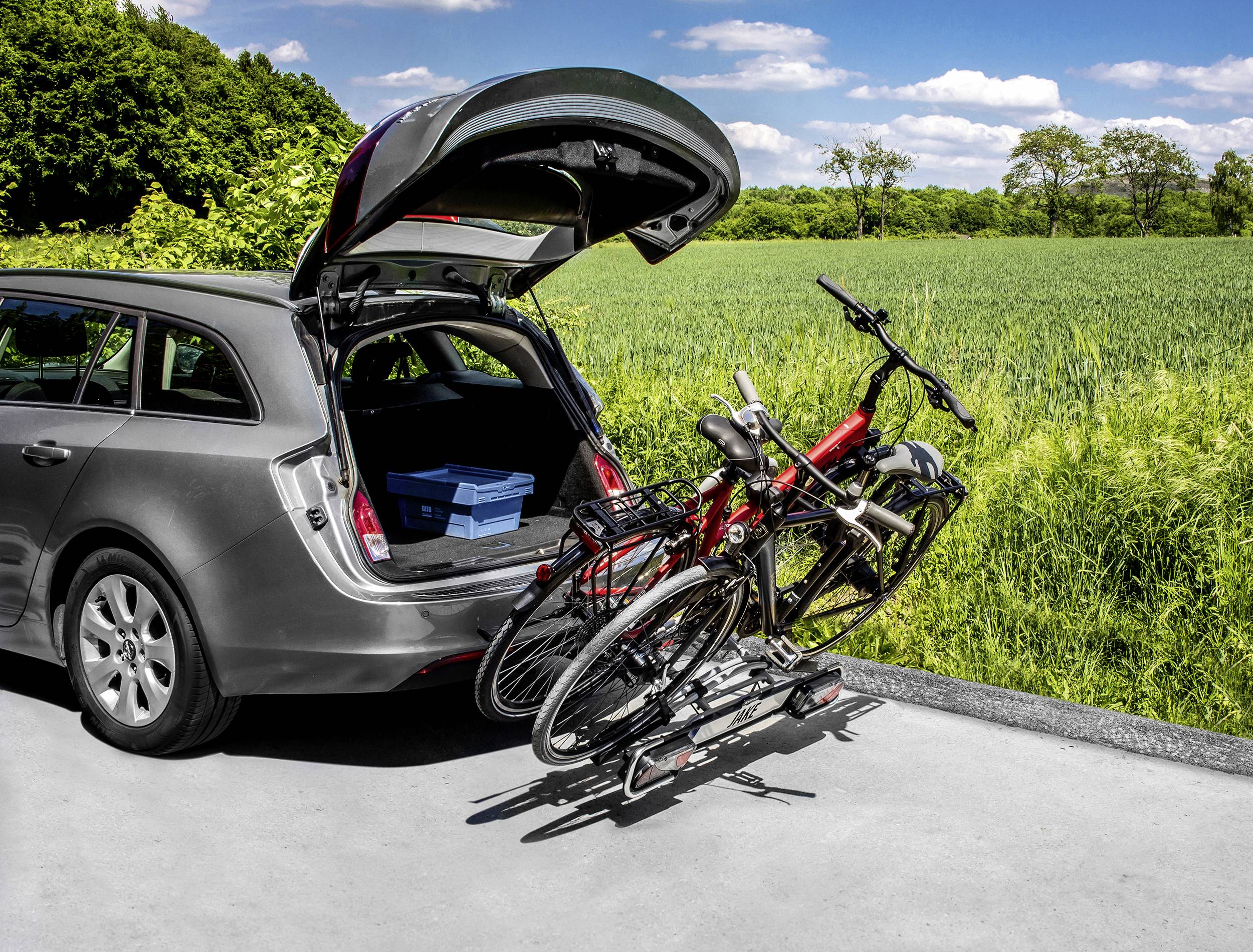 Ein Auto steht am Straßenrand mit geöffneter Heckklappe, auf der ein Fahrradträger mit zwei Fahrrädern montiert ist. Im Hintergrund ist eine grüne Wiese.