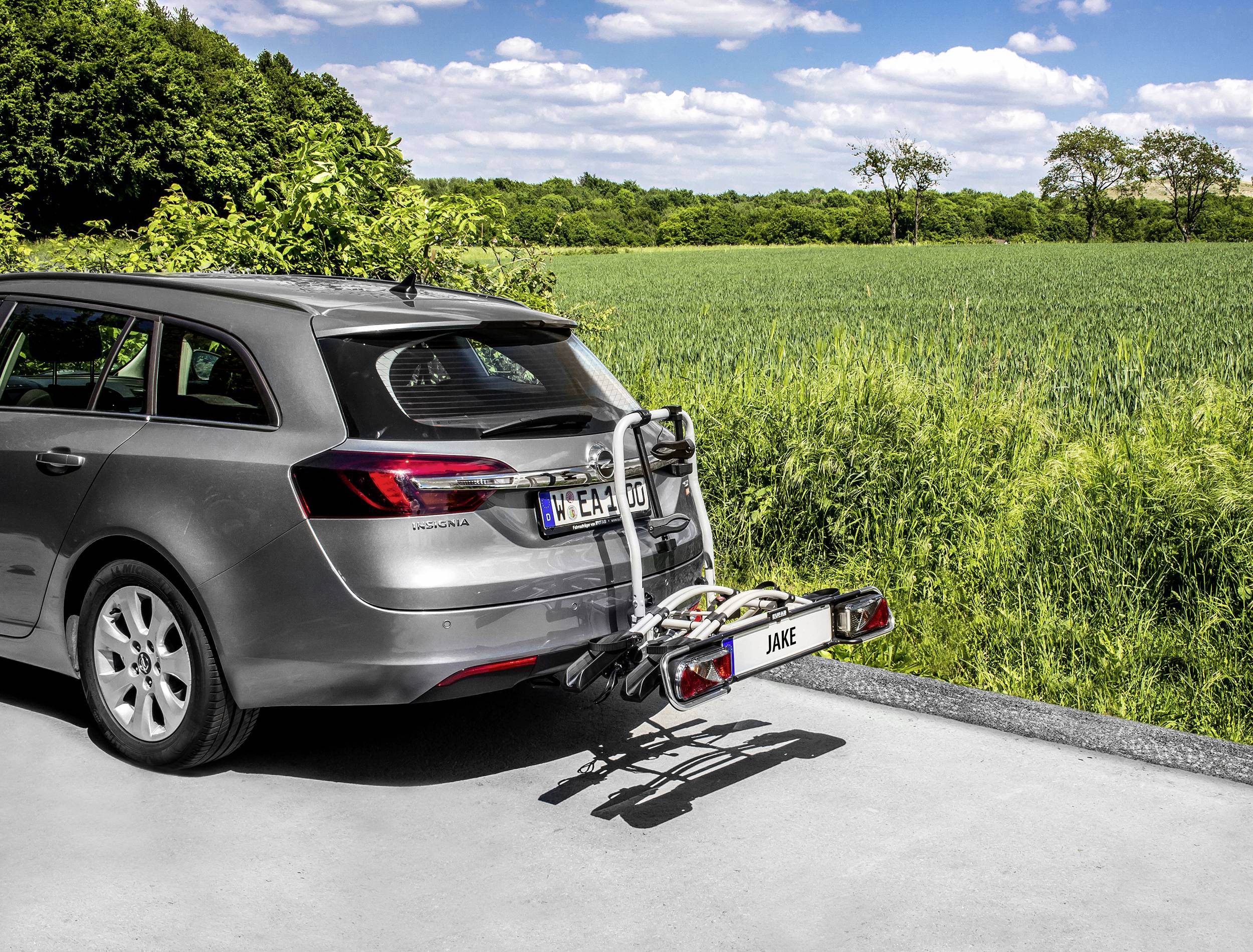 Ein graues Auto mit einem Fahrradträger am Heck steht auf einer Straße vor einem grünen Feld und einem blauen Himmel.