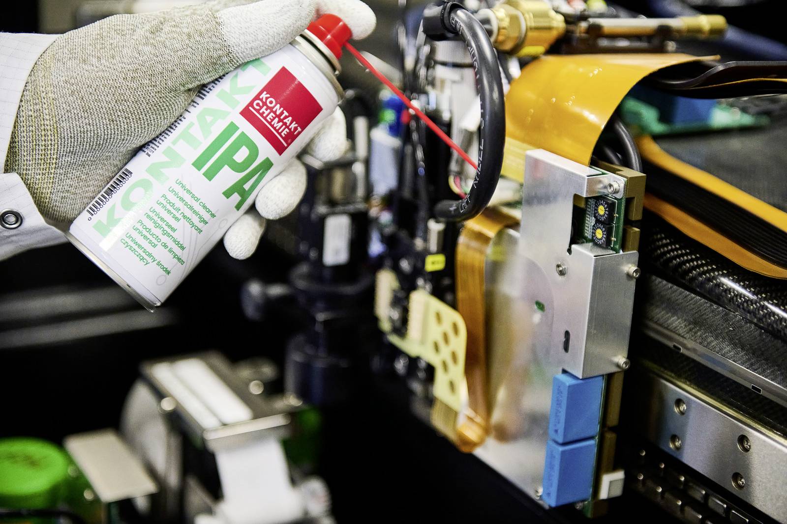 Ein Handschuh hält eine Spraydose mit der Aufschrift 'Kontaktreiniger IPA', während feine Elektronikbauteile in Nahaufnahme sichtbar sind.