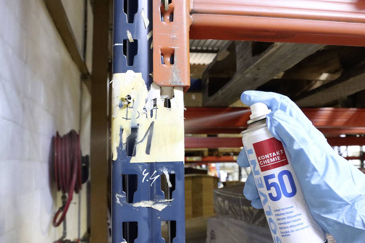 Eine Person sprüht mit blauen Handschuhen ein Lackspray auf ein Metallregal in einem Lager, um Schäden zu reparieren.