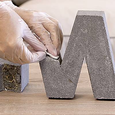 Eine behandschuhte Hand formt sorgfältig eine graue, betonähnliche Skulptur und glättet ihre Oberfläche mit einem Werkzeug, was einen Fokus auf Handwerkskunst andeutet.