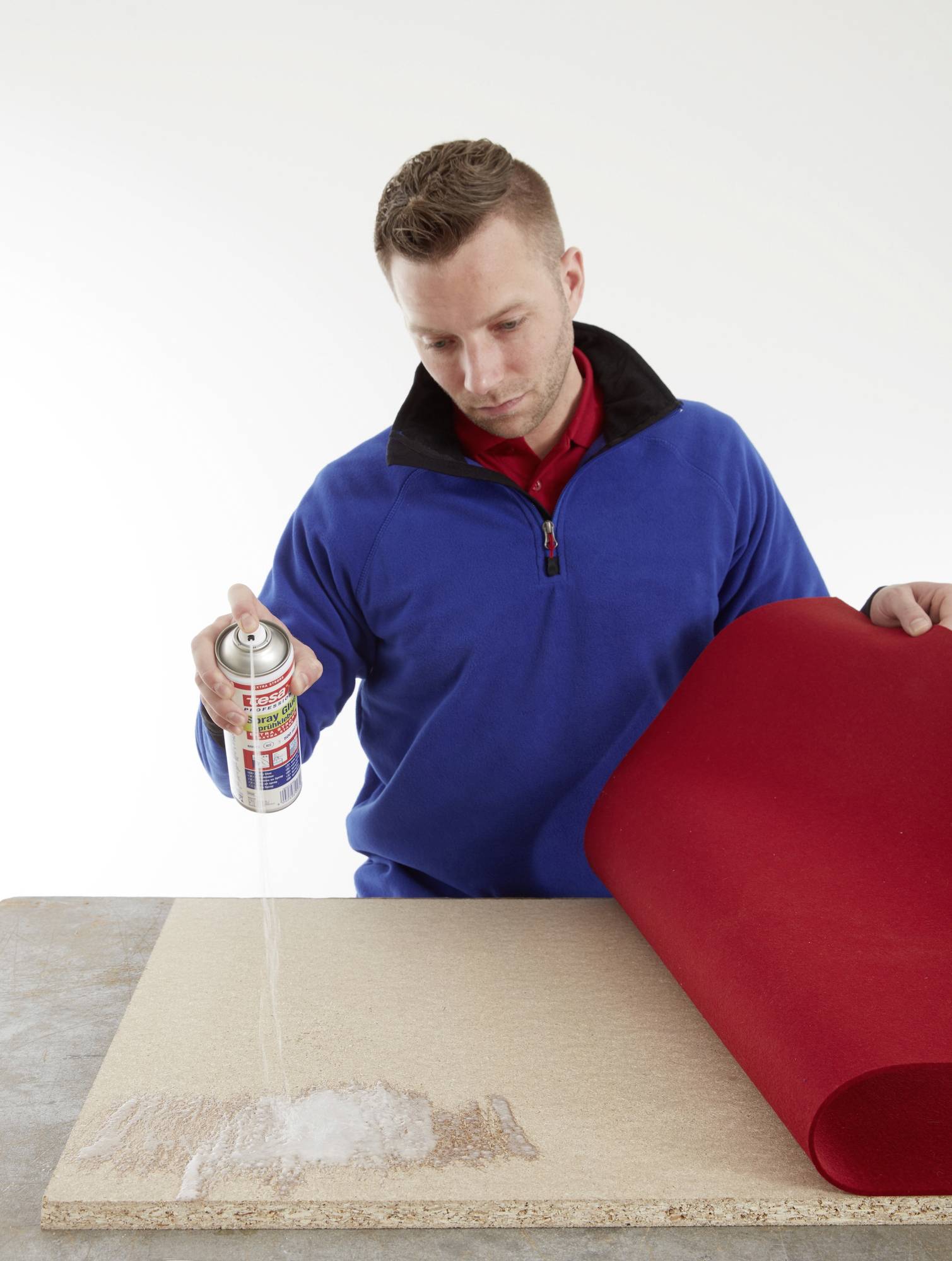 Ein Mann sprüht Kleber aus einer Dose auf ein Holzbrett, während er ein rotes Stoffstück hält, bereit, es darauf zu befestigen.