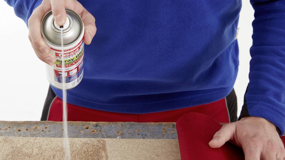 Person sprüht Kleber aus einer Dose auf eine Fläche, während sie ein rotes Stück Stoff hält.