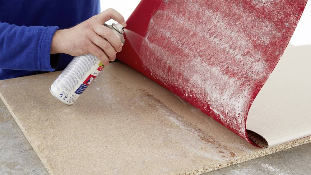 Eine Hand sprüht Kleber aus einer Dose auf ein Stück Holz, während ein rotes Stück Material darauf gelegt wird.