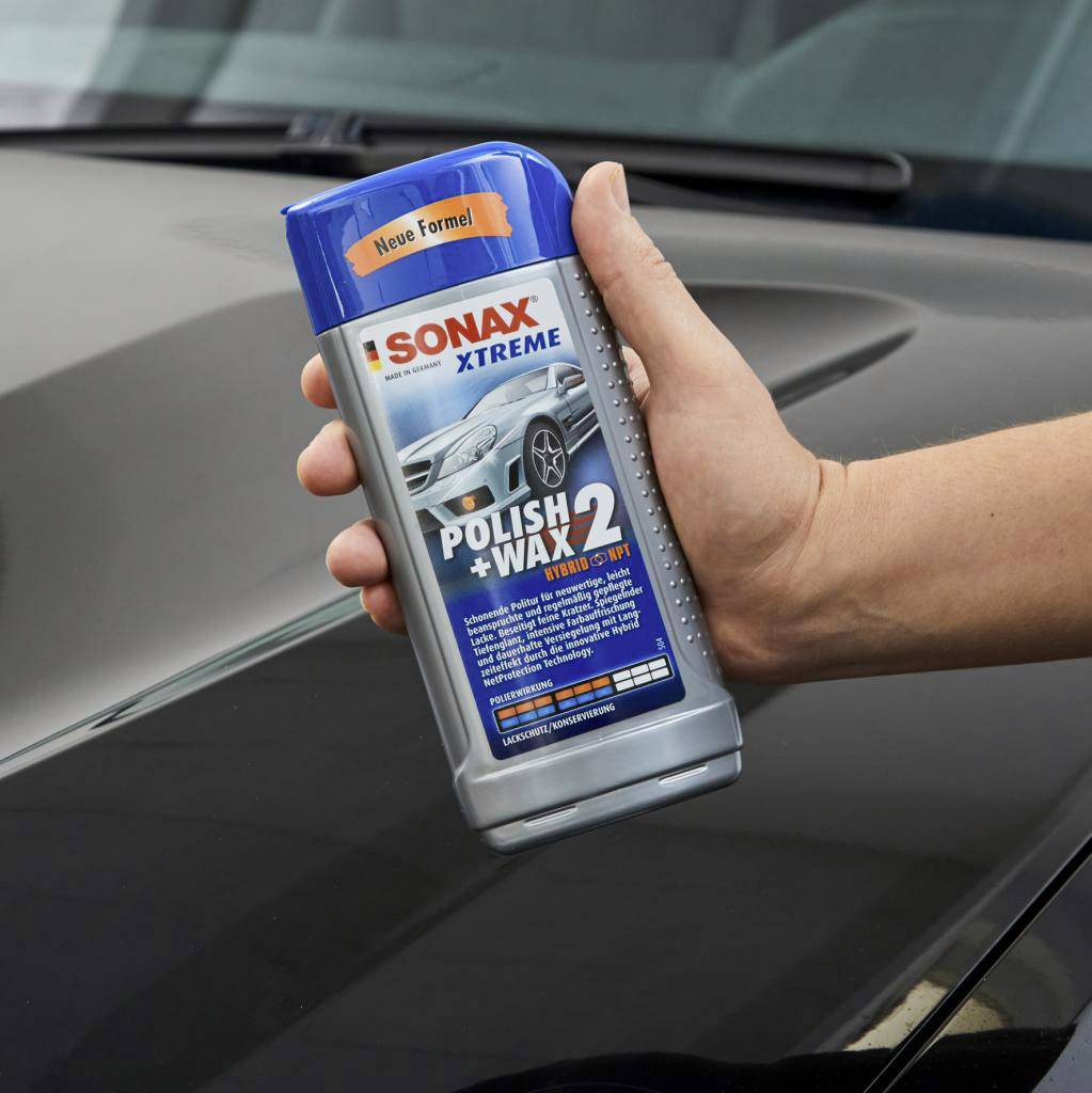 Eine Hand hält eine Flasche mit Autopolitur 'Sonax Xtreme Polish+Wax 2'. Die Flasche hat einen blauen Aufkleber 'Neue Formel'.