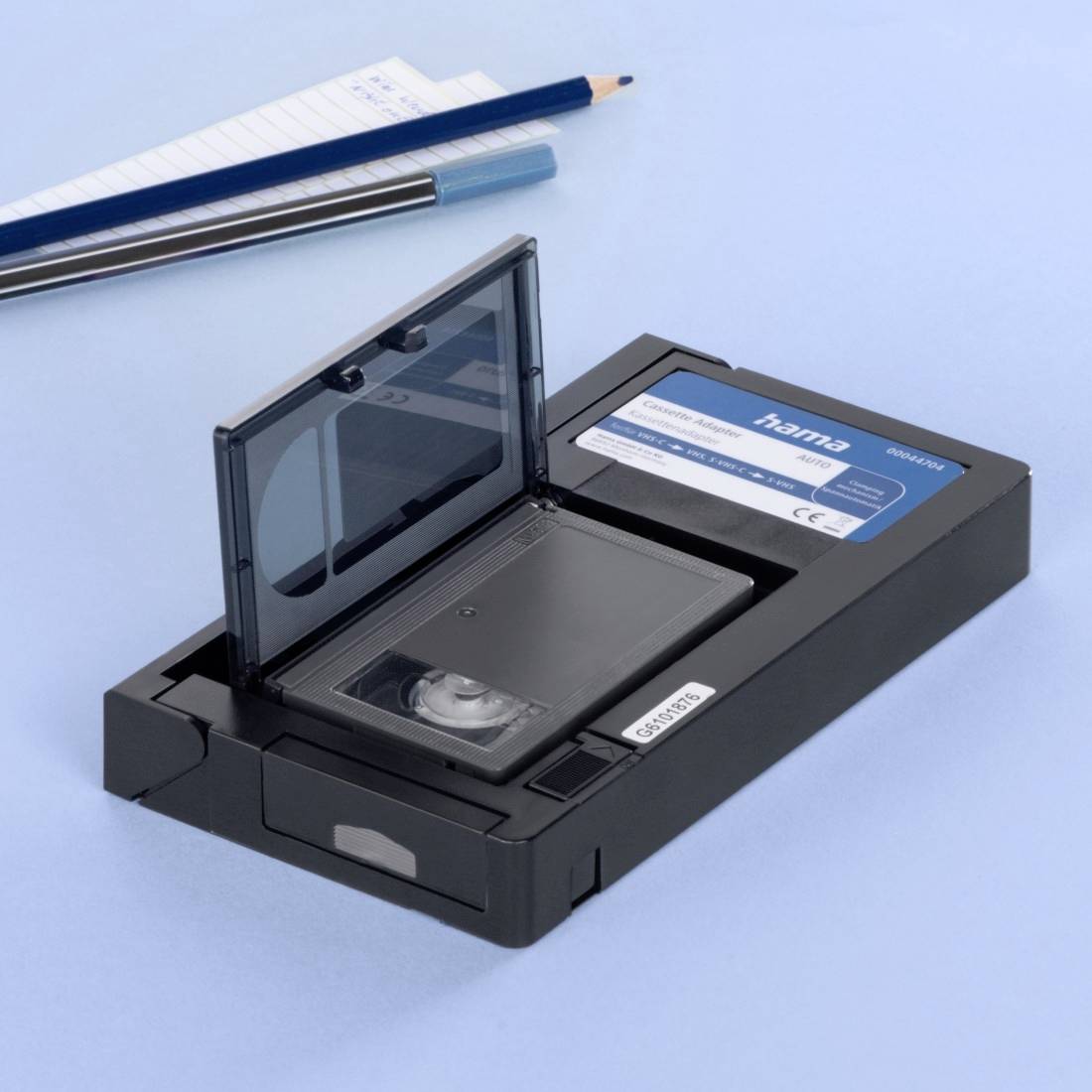 Eine geöffnete VHS-Kassette mit einer offenen Klappe liegt neben einem Notizblock und Stiften auf blauem Hintergrund.