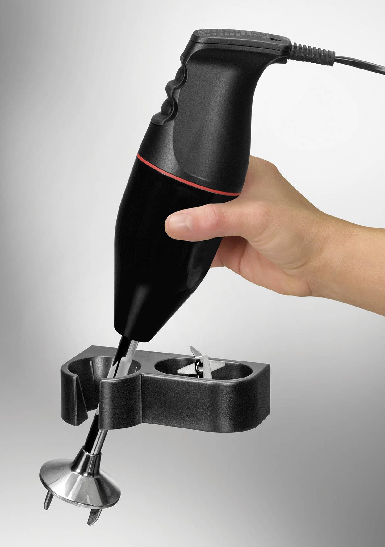 Hand hält einen schwarzen Stabmixer mit Befestigungshalterung, Mixer hat einen edelstahlfarbenen Aufsatz für das Mixen von Zutaten.