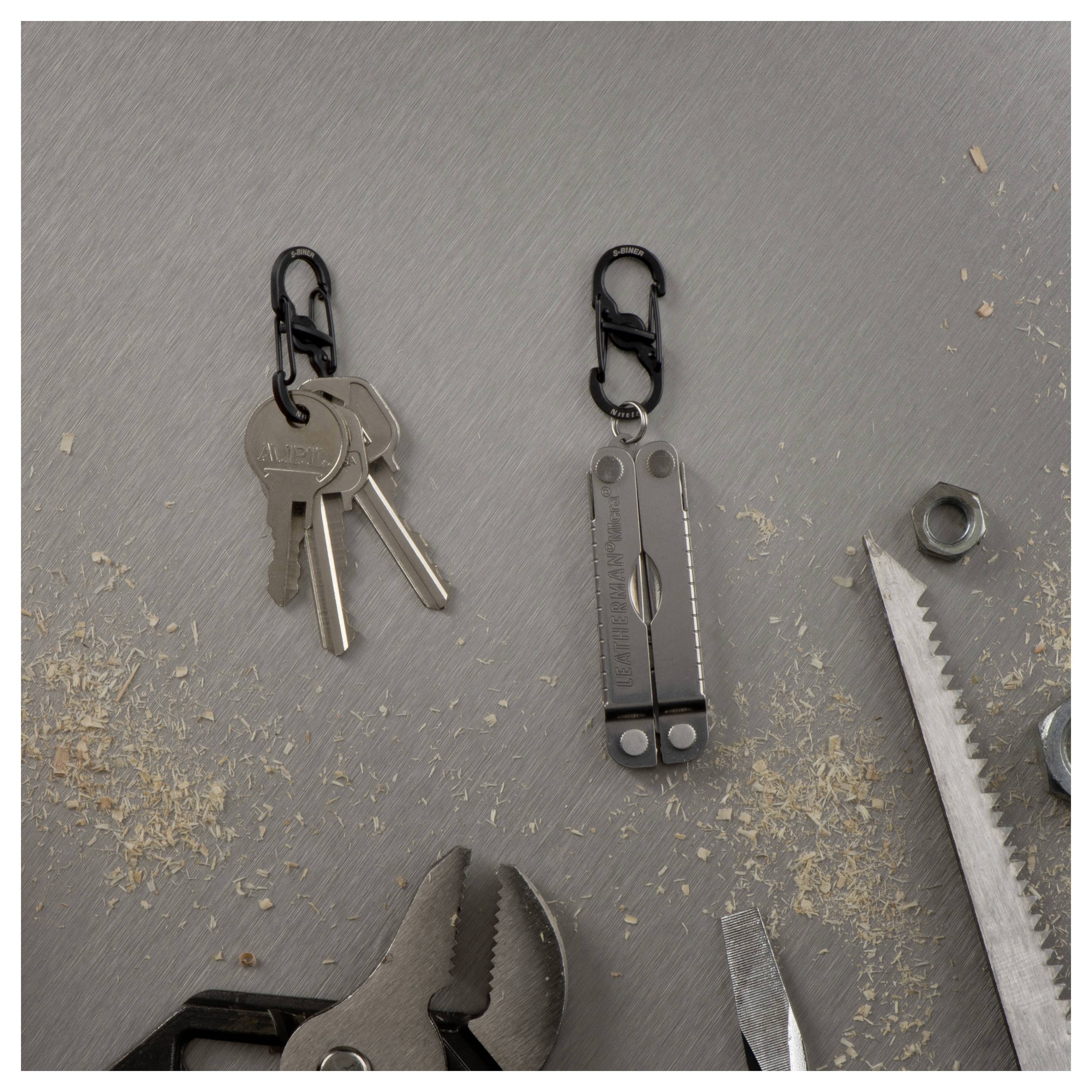 Metalloberfläche mit zwei Schlüsselanhängerhaken; einer hält drei Schlüssel, der andere ein Multitool. Darum herum liegen Sechskantmuttern, ein Schraubenschlüssel und Holzspäne.