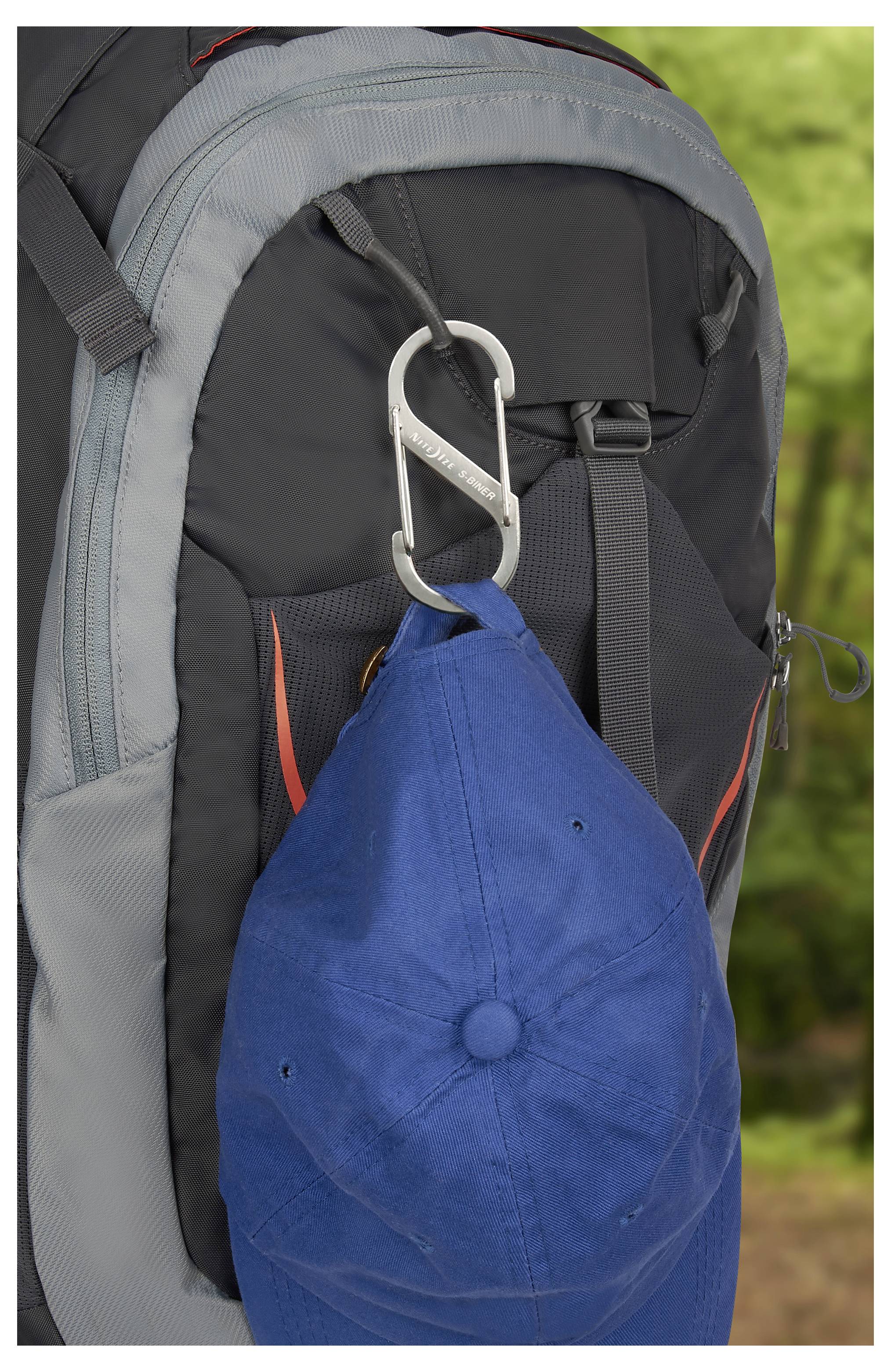 Eine blaue Kappe hängt an einem Karabiner, der an einem schwarzen und grauen Rucksack befestigt ist, mit einem verschwommenen grünen Hintergrund, der eine Outdoor-Umgebung andeutet.