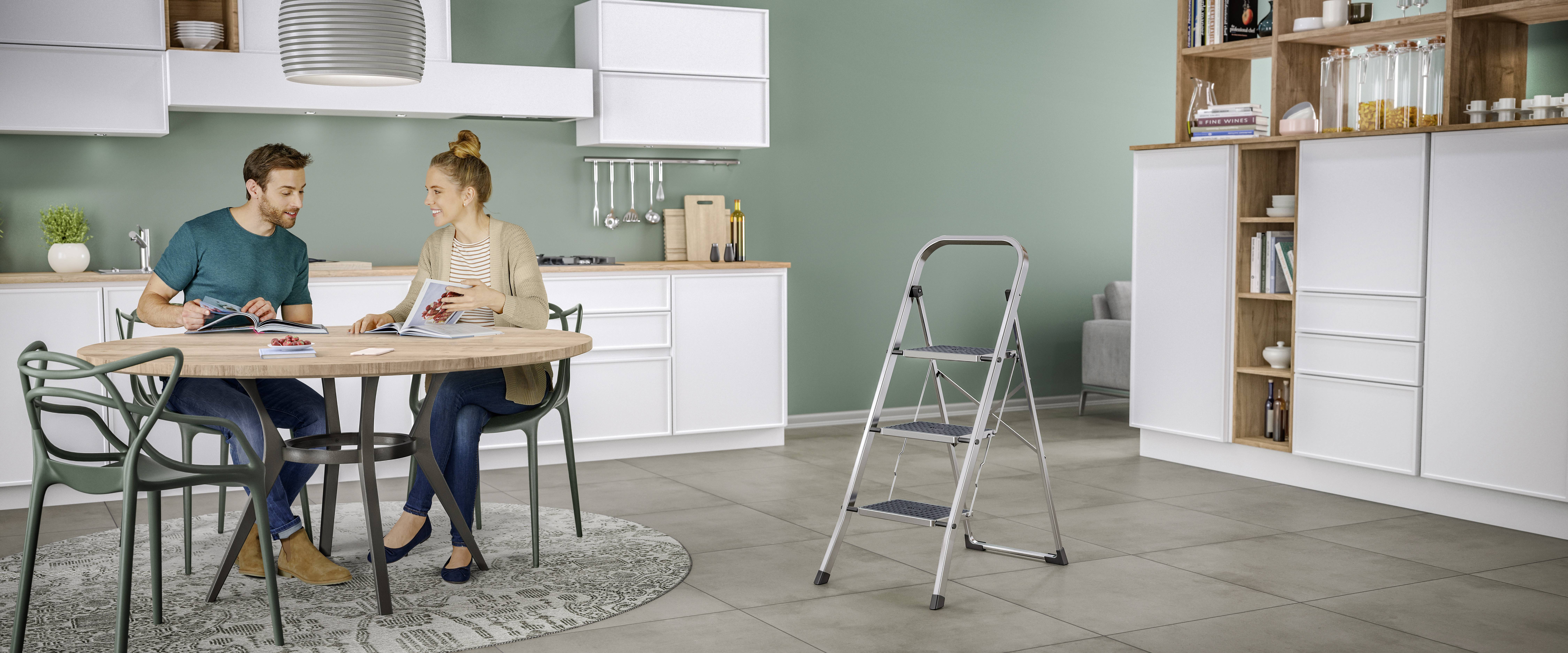 Ein Mann und eine Frau sitzen in einer modernen Küche an einem Tisch und betrachten Farbproben. Eine Trittleiter steht in der Nähe.