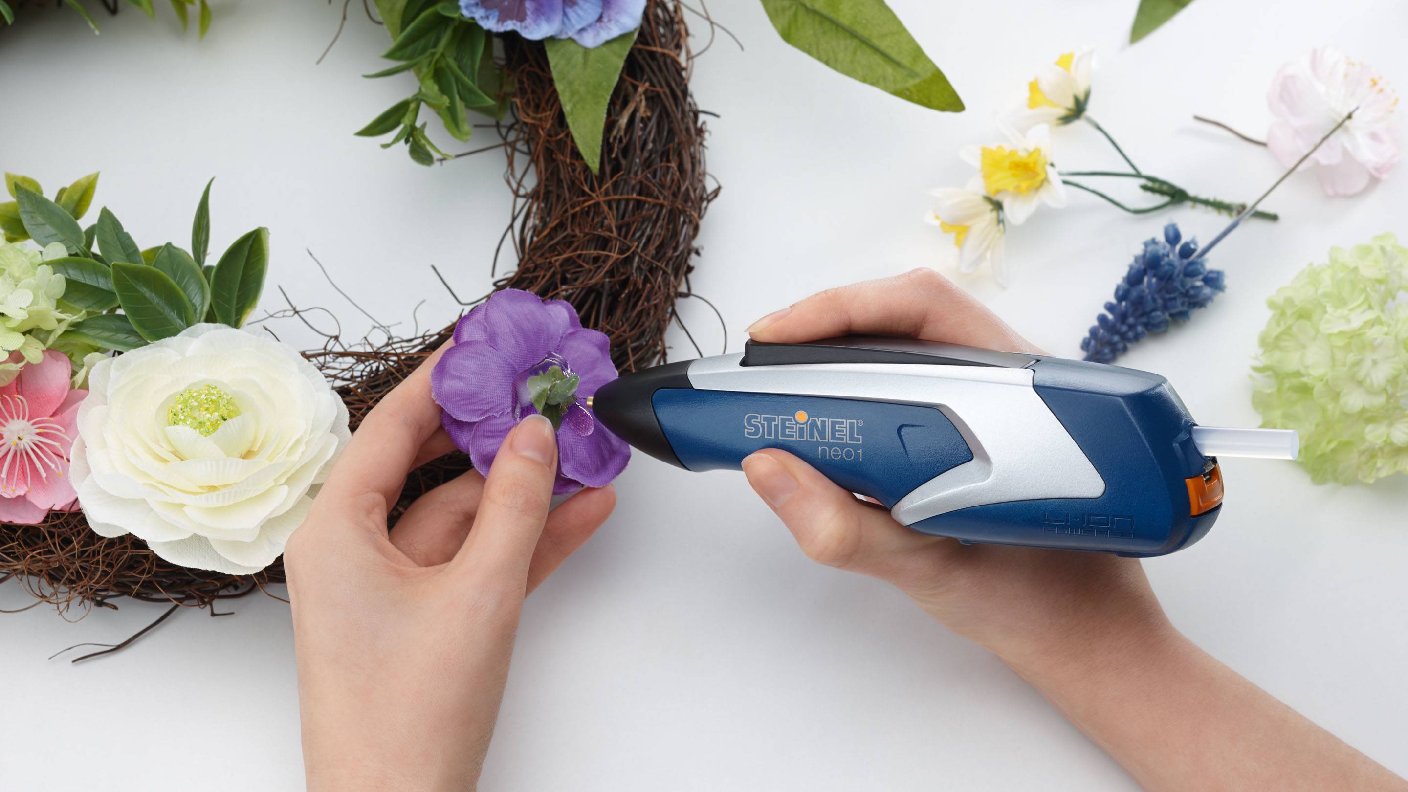 Eine Person klebt eine violette Blume mit einer Heißklebepistole auf einen Kranz aus Naturmaterialien, umgeben von anderen Blumen.
