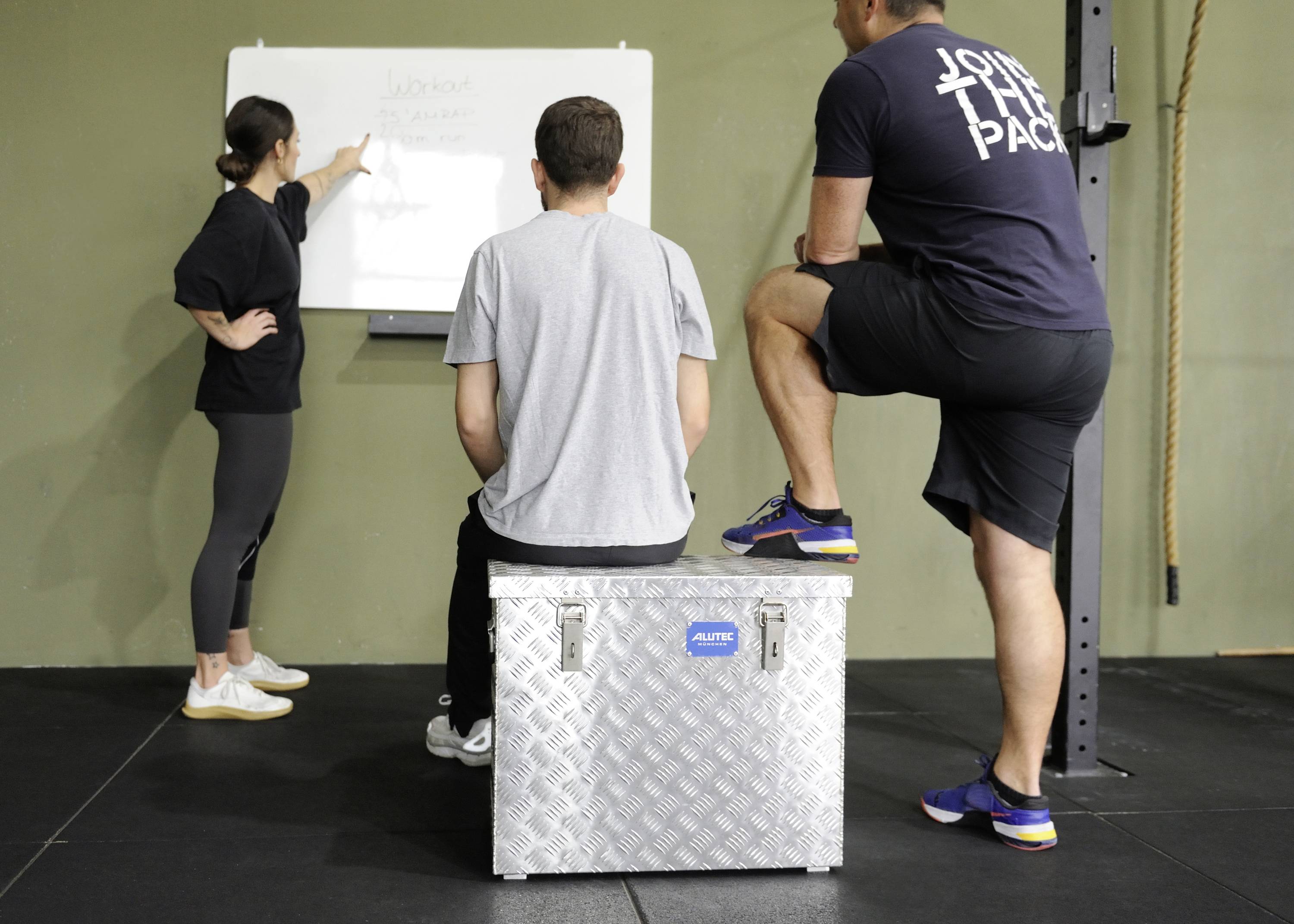 Drei Personen in sportlicher Kleidung diskutieren vor einem Whiteboard in einem Fitnessstudio. Eine Person zeigt auf das Board.