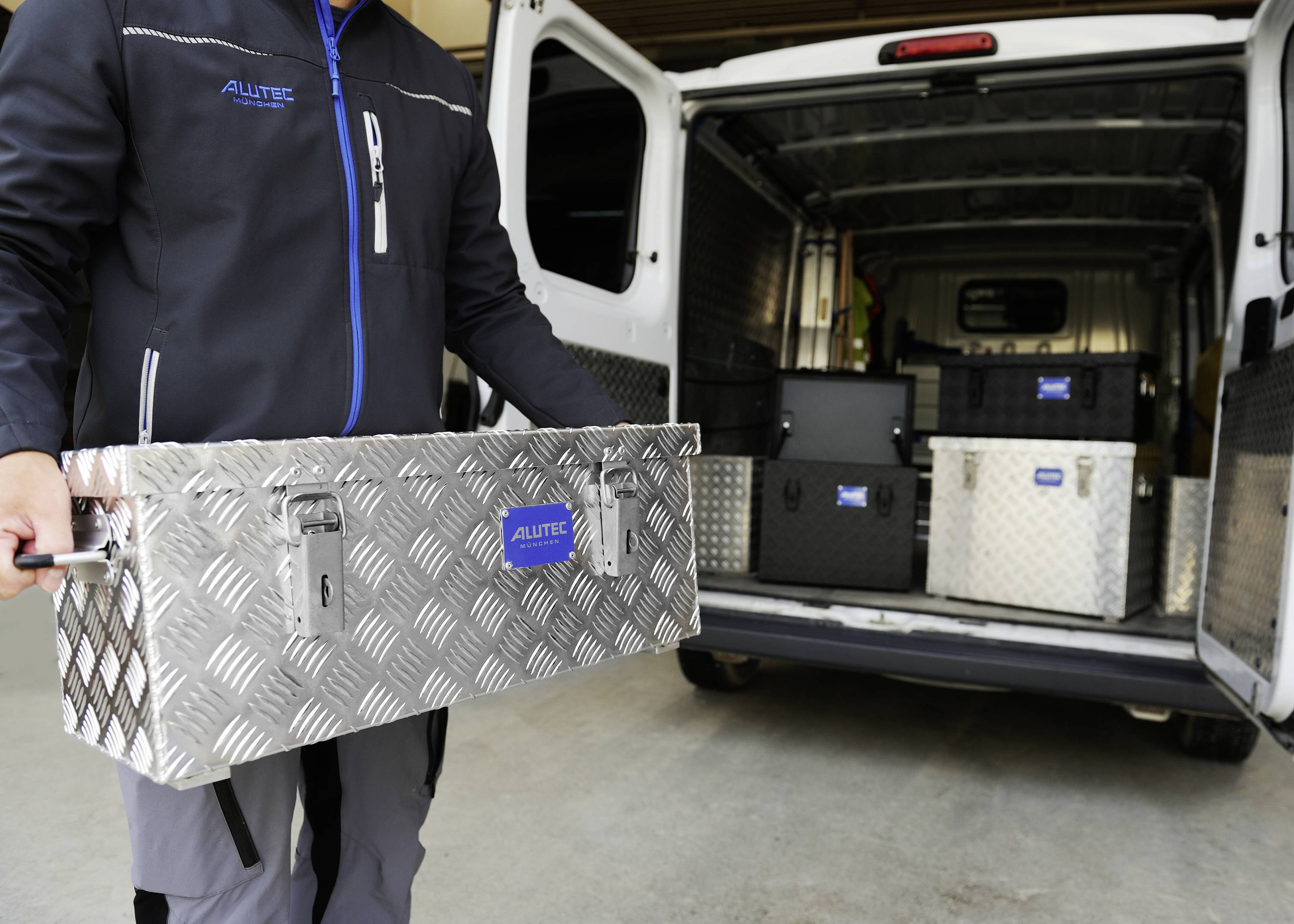 Person lädt Metallboxen mit Griffen in einen Lieferwagen. Die Boxen sind aus Riffelblech und tragen das Logo 'ALUTEC'.