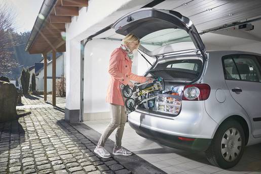 Person packt Gegenstände in den geöffneten Kofferraum eines Autos in einer Garage. Der Morgen scheint sonnig und ruhig zu sein.