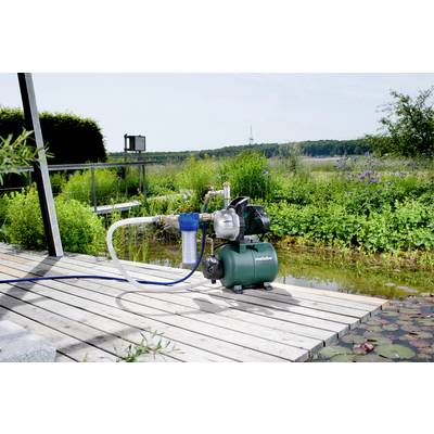 Eine Wasserpumpe auf einer Holzterrasse vor einem Teich, umgeben von Pflanzen. Im Hintergrund ist eine Landschaft mit Bäumen sichtbar.