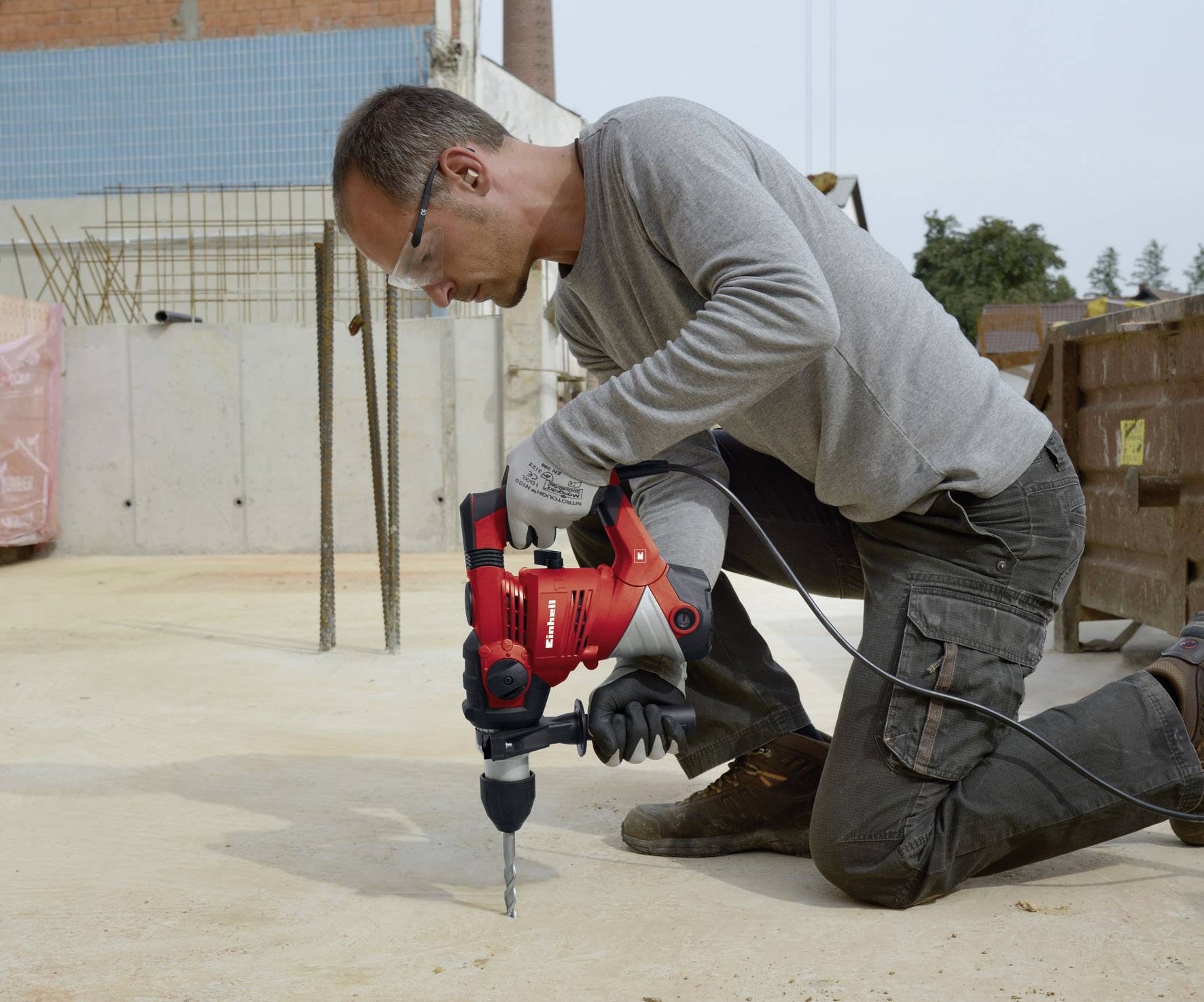 Ein Mann bohrt auf einer Baustelle mit einem roten Bohrer in den Betonboden, trägt Schutzkleidung und kniet auf dem Boden.
