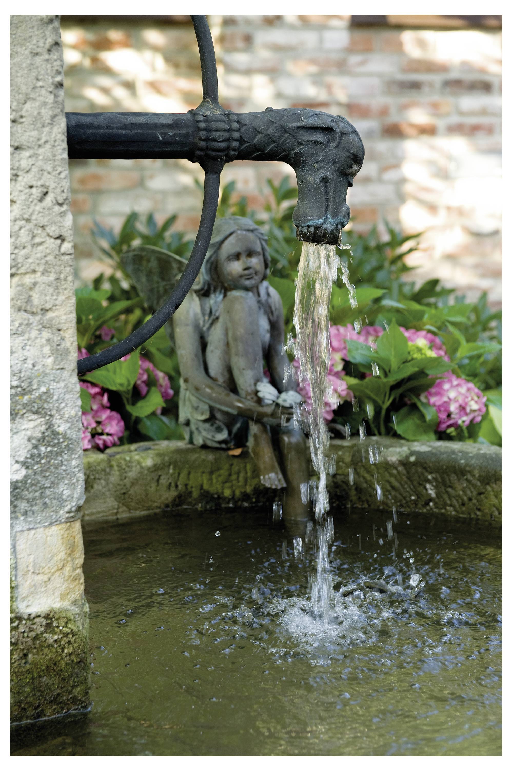 Ein Steinbrunnen mit einem drachenartig geformten Auslauf gießt Wasser in einen Teich, umgeben von üppiger Vegetation und rosa Blumen, mit einer Feenstatue.