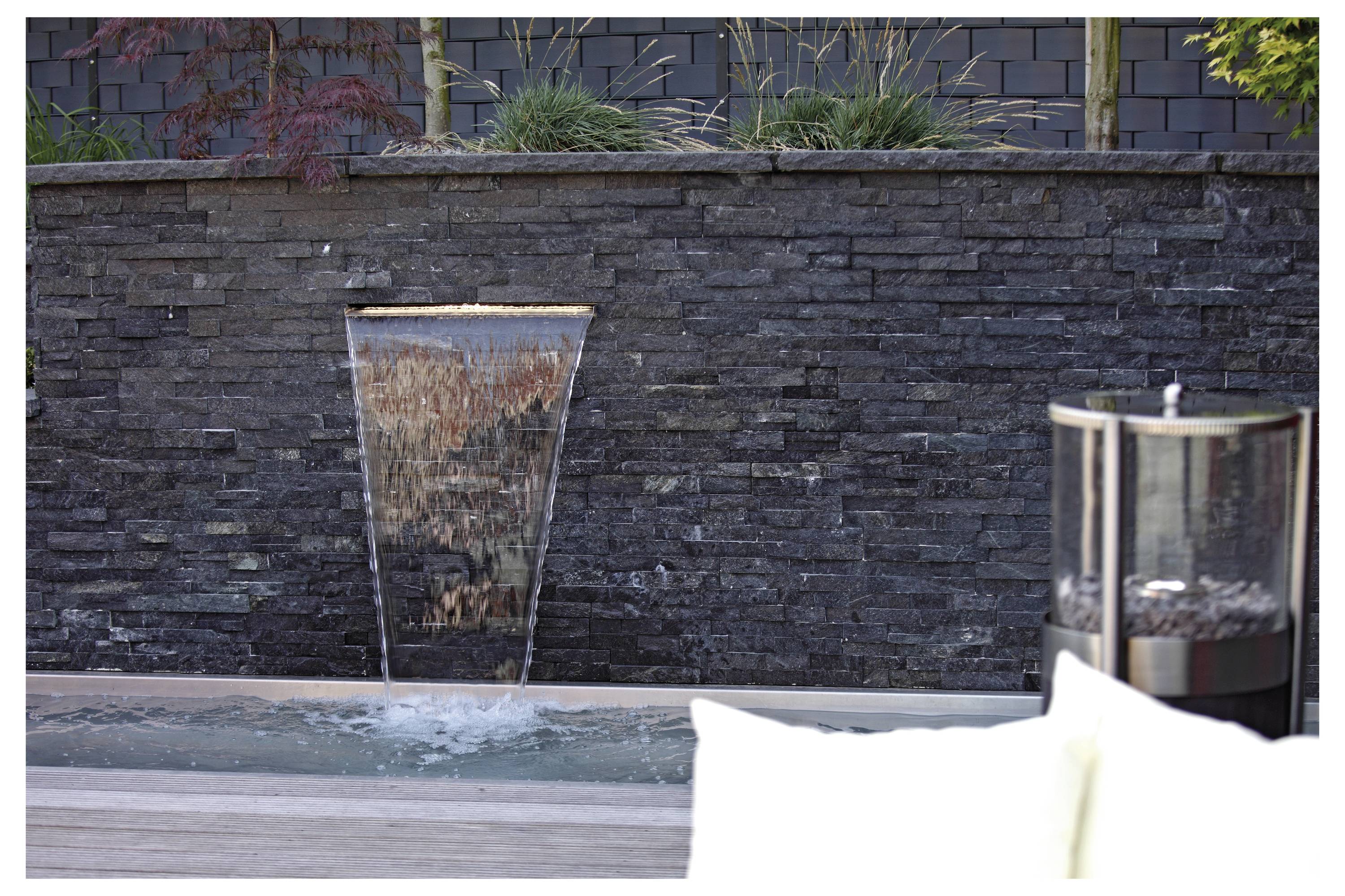 Ein moderner Garten mit einer Steinmauer zeigt einen Wasserfall, der in einen rechteckigen Pool herunterstürzt.