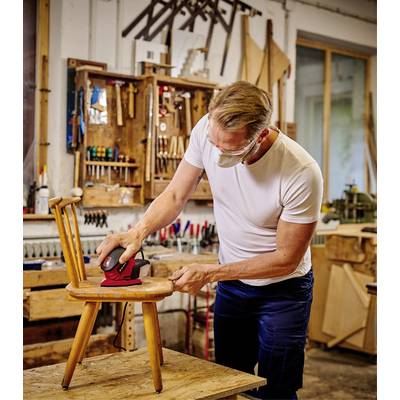 Eine Person schleift in einer Werkstatt einen Holzstuhl mit einer Schleifmaschine. Im Hintergrund sind Werkzeuge und Holz zu sehen.