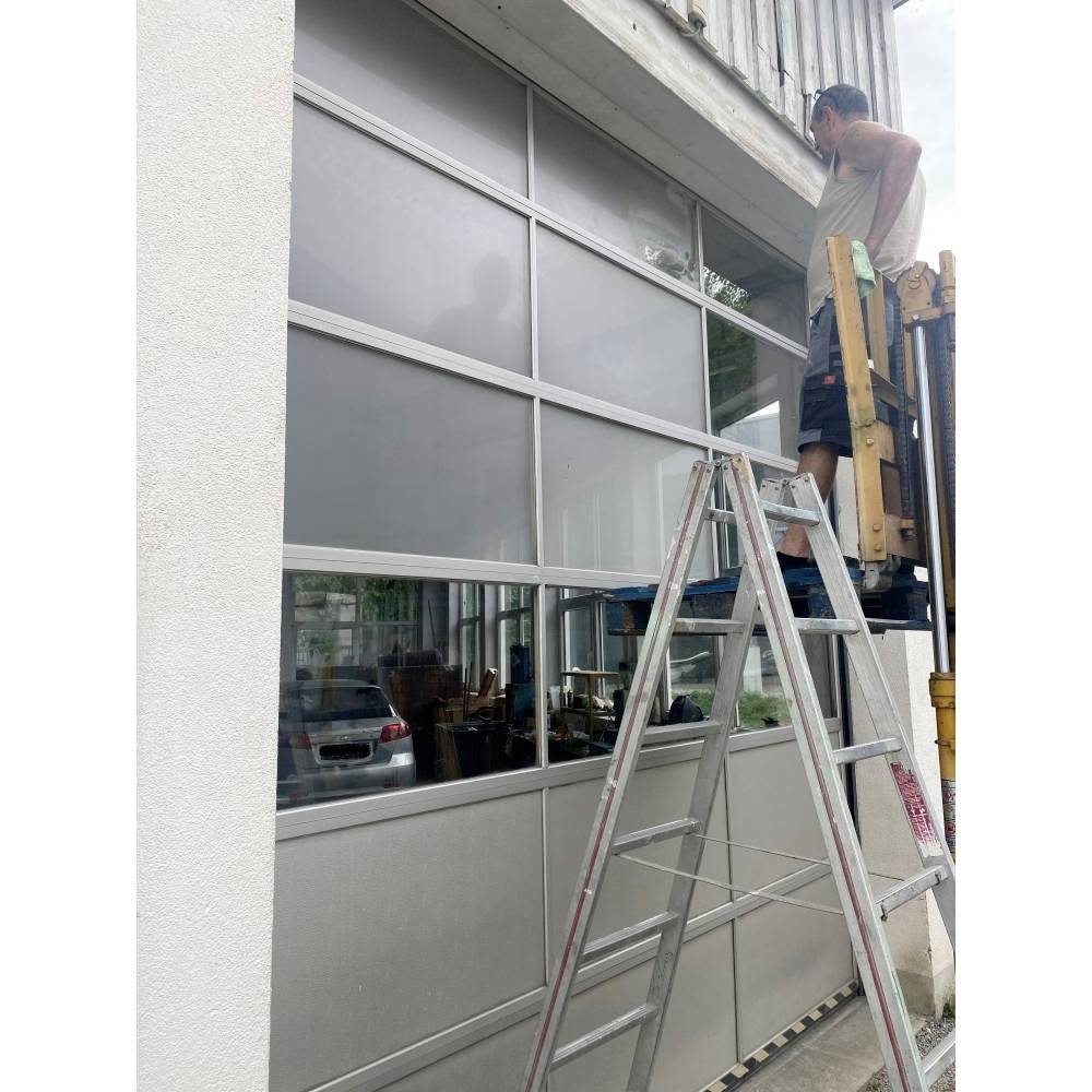 Ein Mann arbeitet von einer Hubarbeitsbühne aus an einem Garagentor, eine Leiter steht daneben. Die Szene deutet auf Wartungs- oder Reparaturarbeiten hin.