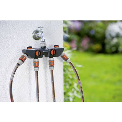Gartenschlauchverteiler mit vier Anschlüssen an einer Außenwand montiert, verbunden mit Schläuchen, umgeben von grünem Rasen und bunten Blumen.