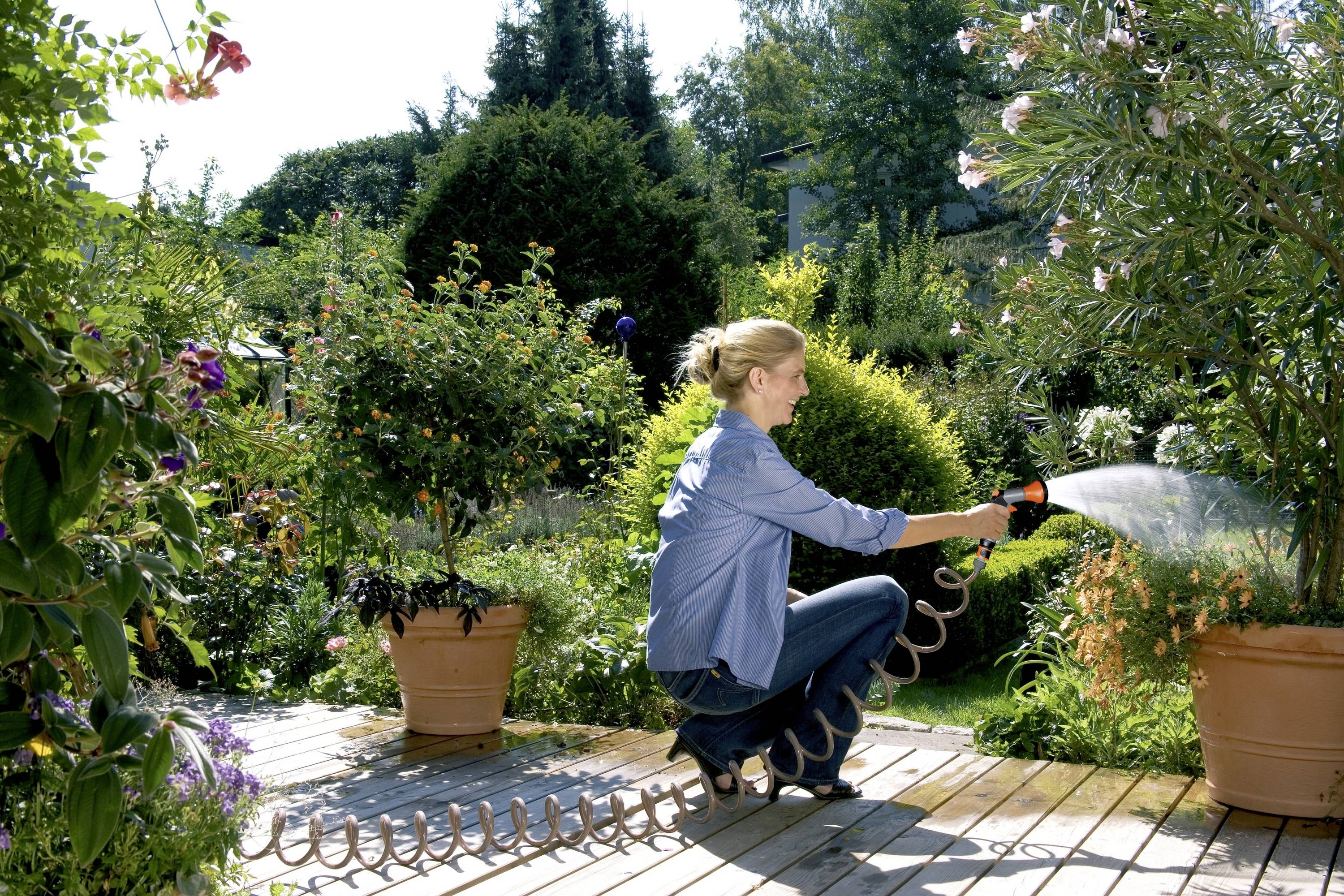 Eine Frau gießt an einem sonnigen Tag einen Garten mit einem Wasserschlauch. Umgeben von blühenden Pflanzen und Topfbäumen.