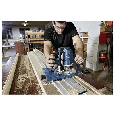 Handwerker nutzt Elektrowerkzeug zum Fräsen in Holzwerkstatt, um Holzleiste zu bearbeiten. Sägespäne auf Tisch, Regale im Hintergrund.