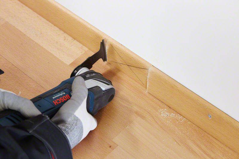 Eine Hand hält ein oszillierendes Werkzeug und schneidet einen Abschnitt einer Holzfußleiste an der Wand.