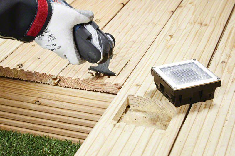 Eine Person bearbeitet mit einem Elektrowerkzeug Holzplanken für eine Terrasse. Eine quadratische Lampe ist in das Holz eingelassen.