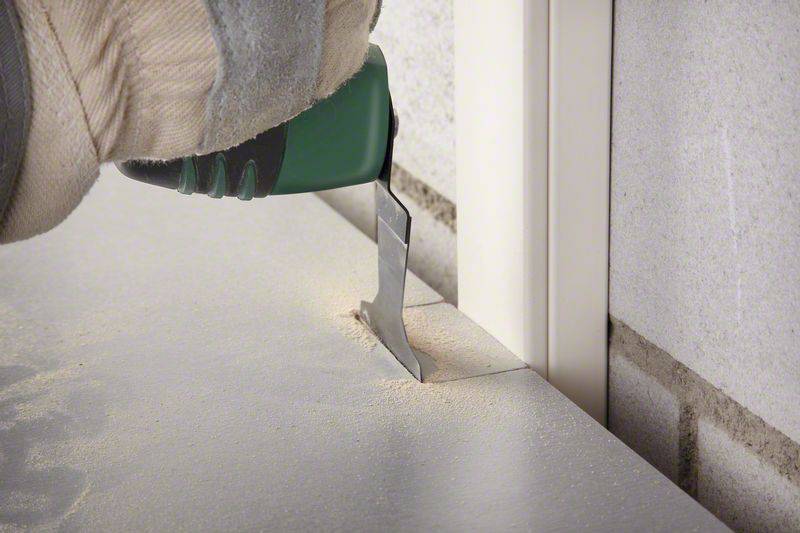 Eine Hand mit Handschuh hält ein multifunktionales Werkzeug, das eine präzise Schnittlinie in einer Oberfläche nahe an einer Wand führt.