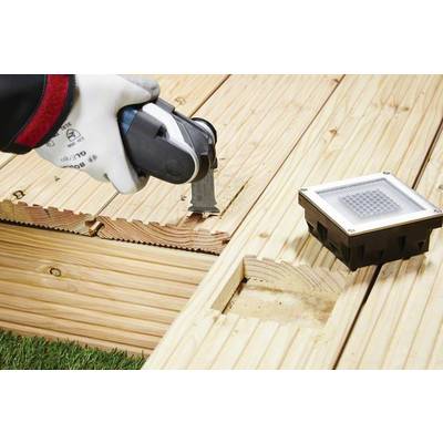 Eine Person montiert eine Terrasse aus Holzdielen mit einem Elektrowerkzeug. Neben der Arbeit liegt eine quadratische Bodenleuchte.