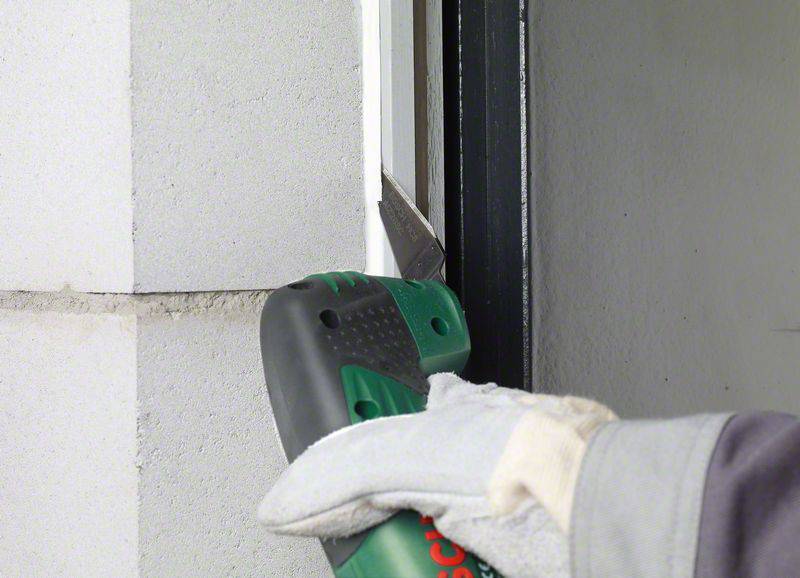'Ein Arbeiter trägt Handschuhe und schneidet mit einem grünen Werkzeug Dichtungsmaterial zwischen einer Wand und einem Metallrahmen ab.'