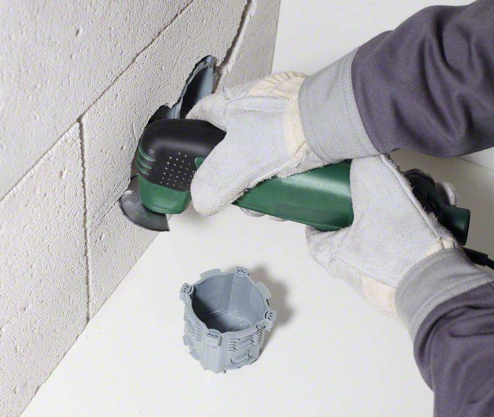 Person schneidet mit einem Elektrowerkzeug ein Loch in eine weiße Wand. Eine graue Steckdose liegt daneben.