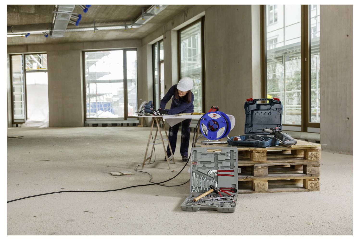 Bauarbeiter in Schutzhelm benutzt Elektrowerkzeug auf einer Werkbank in einem unfertigen Gebäude, mit Werkzeugen und Kabeln in der Nähe.
