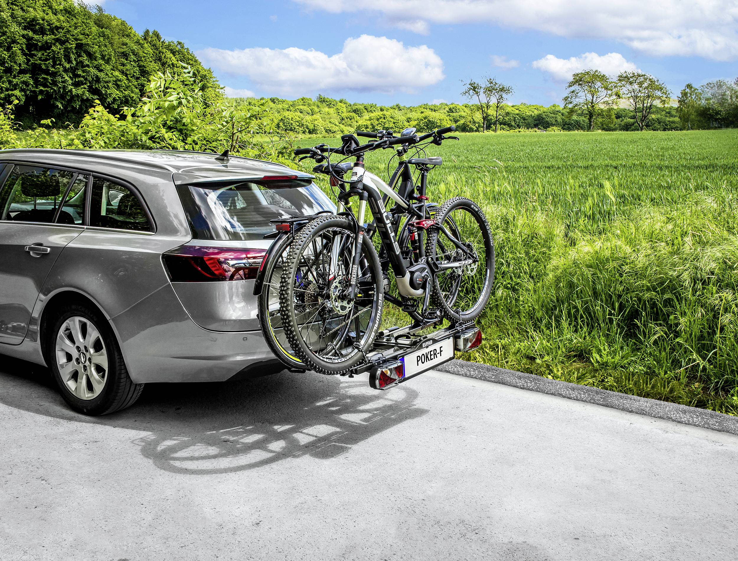 Ein silberner Kombi mit zwei Fahrrädern auf einem Heckträger steht am Straßenrand. Im Hintergrund ist eine grüne Wiese zu sehen.