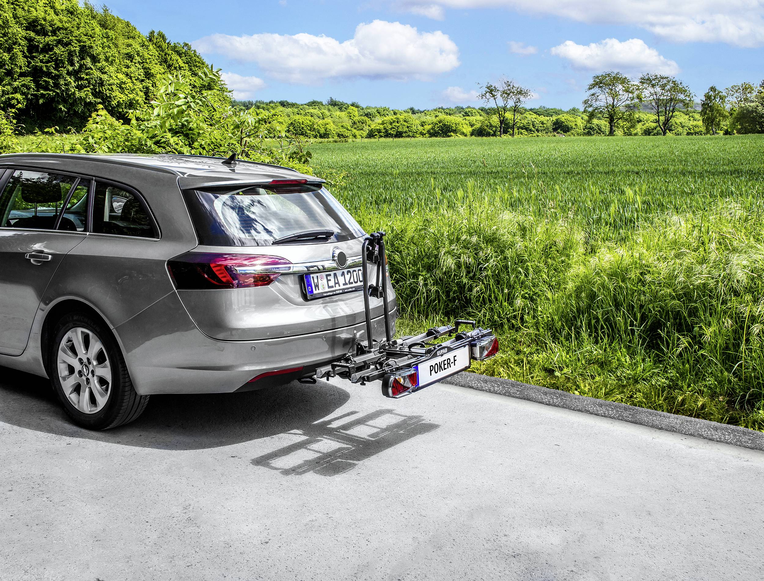 Ein silbergrauer Kombi mit einem montierten Fahrradträger auf einem Landweg vor einem grünen Feld und bewölktem Himmel.