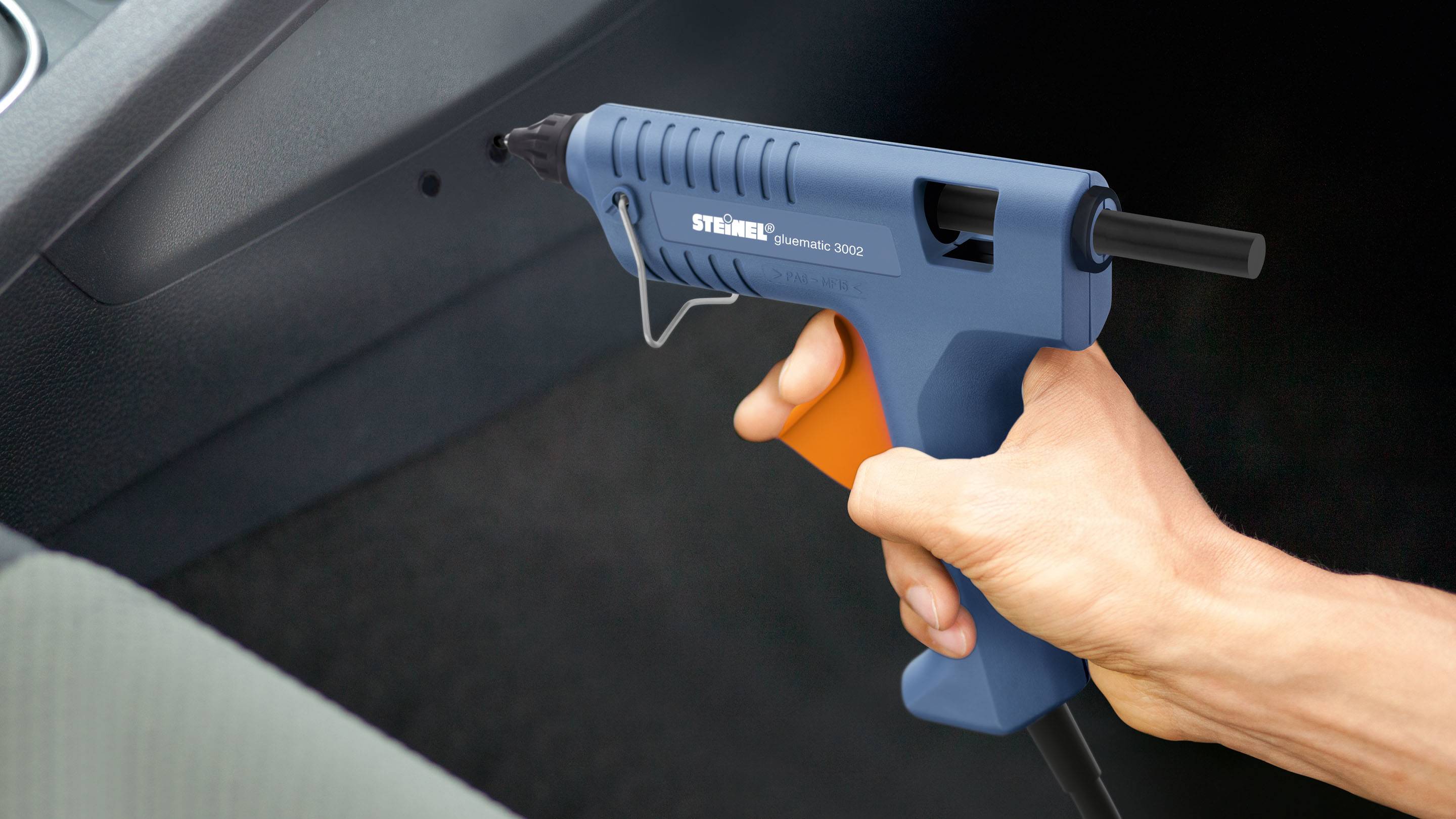 Eine Person hält eine blaue Heißklebepistole in der Hand, bereit zum Einsatz. Die Umgebung deutet auf eine Werkstatt oder Hobbyraum hin.