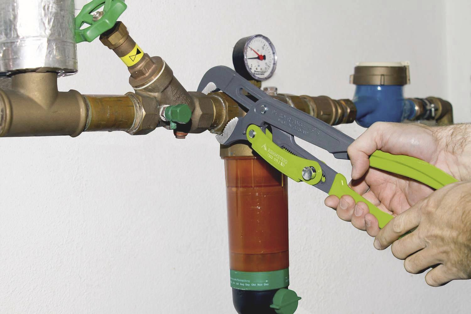 Eine Person zieht eine Rohrzange an einem Ventil eines Kupferrohrs an; im Hintergrund ein Manometer sowie ein blaues Bauteil.