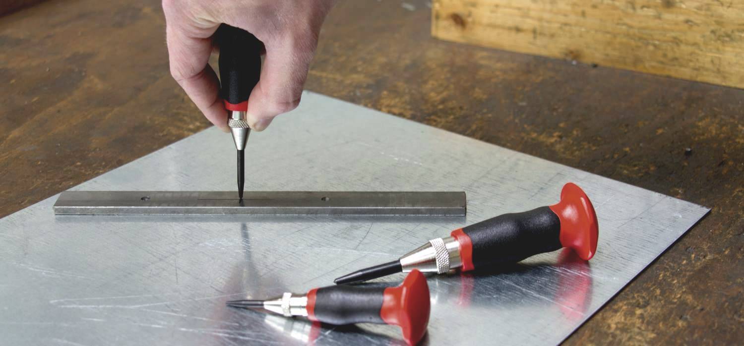 Hand hält einen Druckkörner über einer Metallplatte, daneben liegen zwei weitere Druckkörner. Hintergrund ist eine Holzoberfläche.