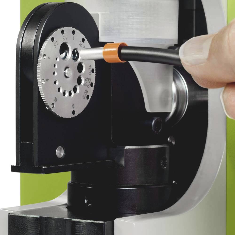 Ein Finger hält einen Faseroptikstecker, der in ein schwarzes, zahnradähnliches Gerät eingesteckt wird, vor einem grünlichen Hintergrund.