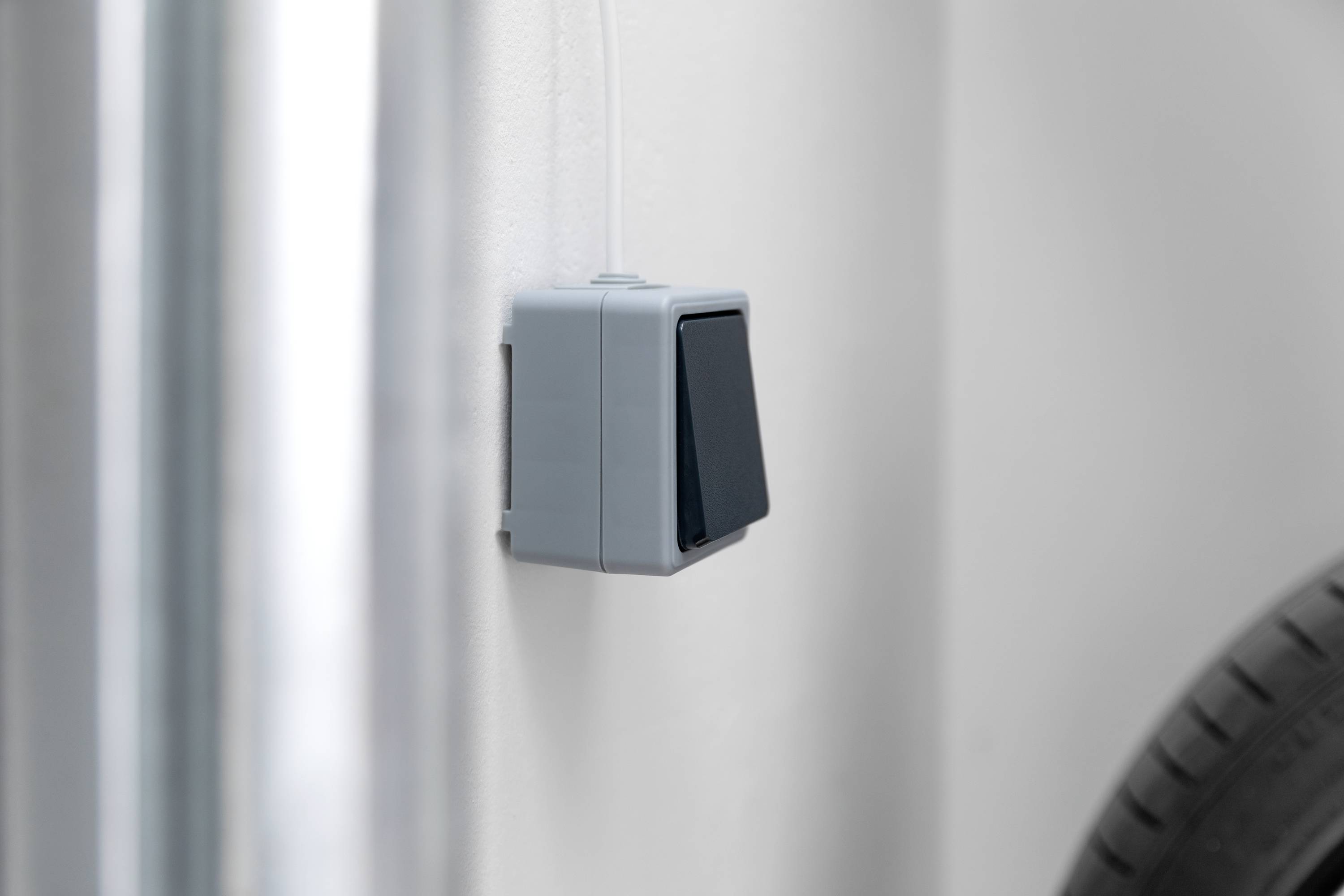 Ein grauer Sensor ist an einer weißen Wand montiert. Im Vordergrund sind verschwommene Details von Rohren und einem Reifen erkennbar.