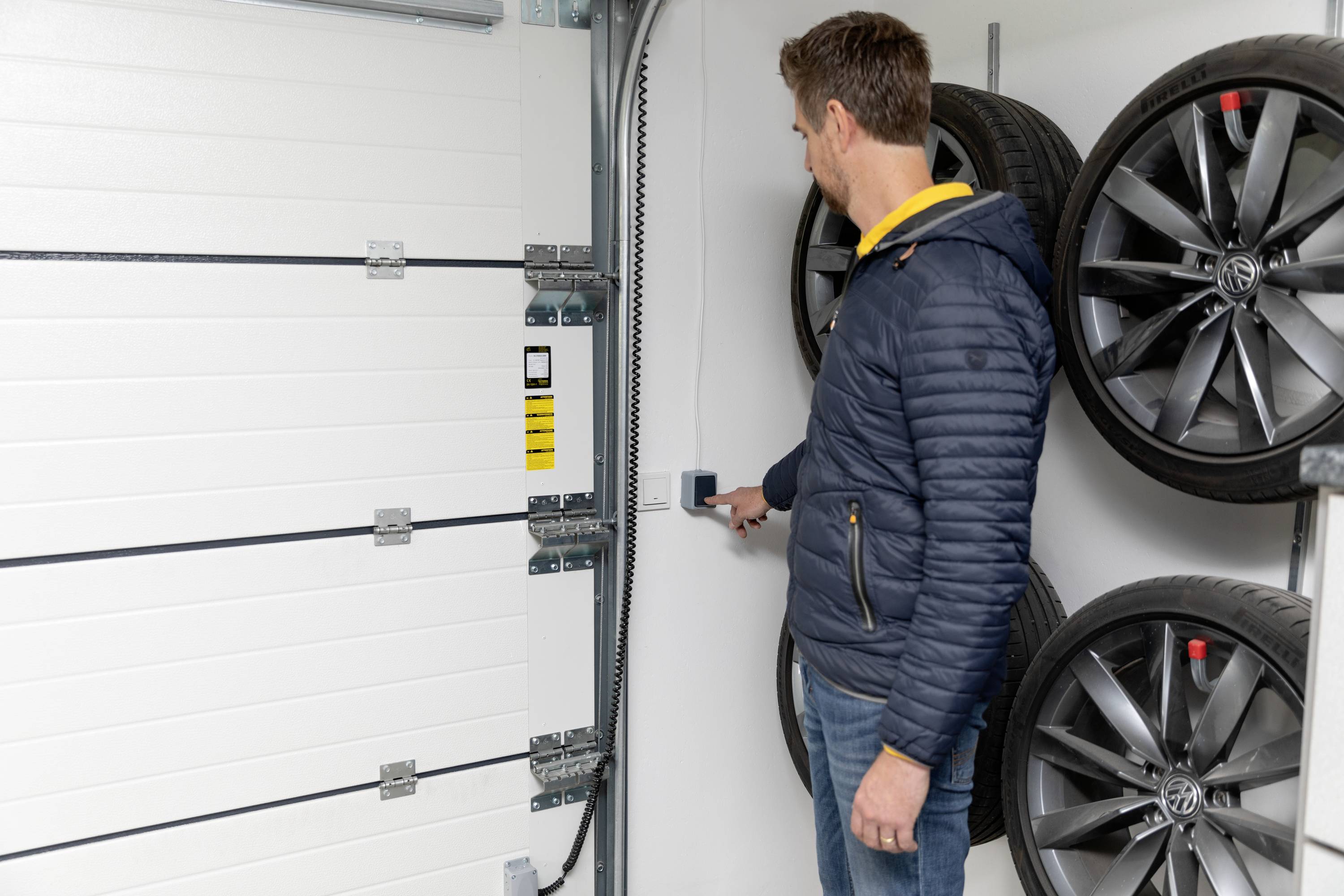 Ein Mann im blauen Anorak öffnet mit einem Wandschalter ein Garagentor. Im Hintergrund sind Autoreifen an der Wand montiert.