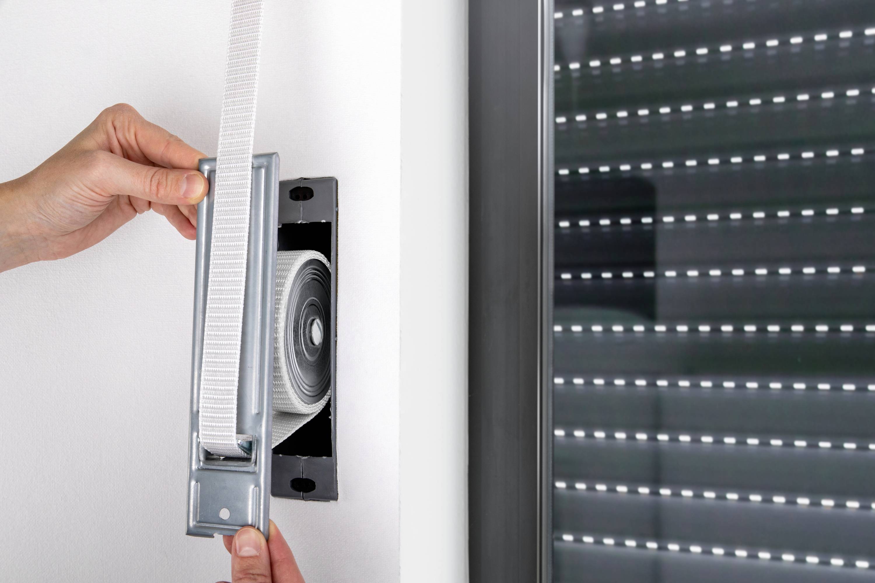 Hände montieren oder justieren einen Rollladengurt an einem Fenster, das teilweise mit einem geschlossenen Rollladen bedeckt ist.
