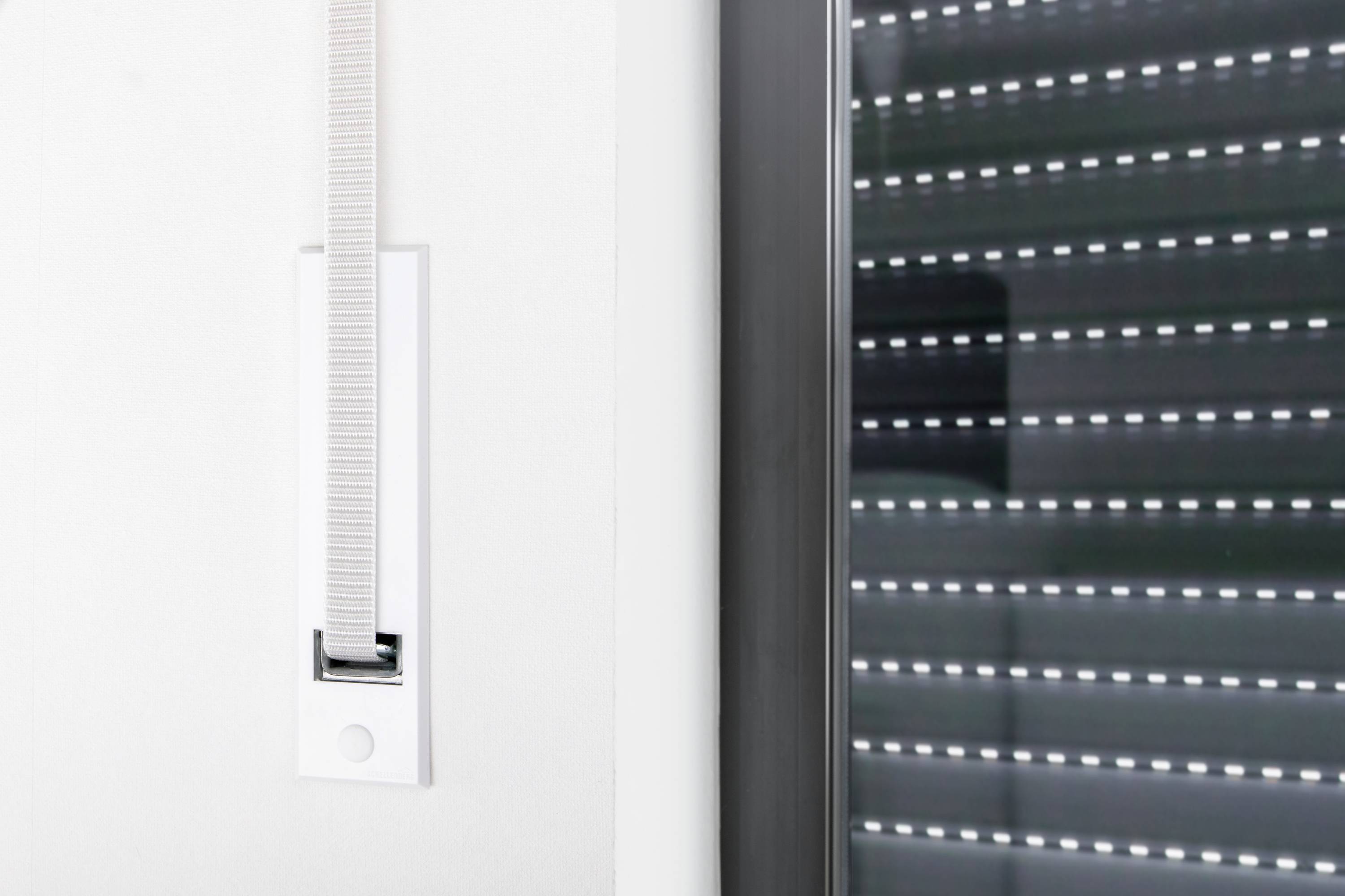 Ein Fenster mit einem geschlossenen Rollladen und einem Gurtwickler an der Wand daneben. Der Rollladen ist aus Metall mit schmalen Schlitzen.