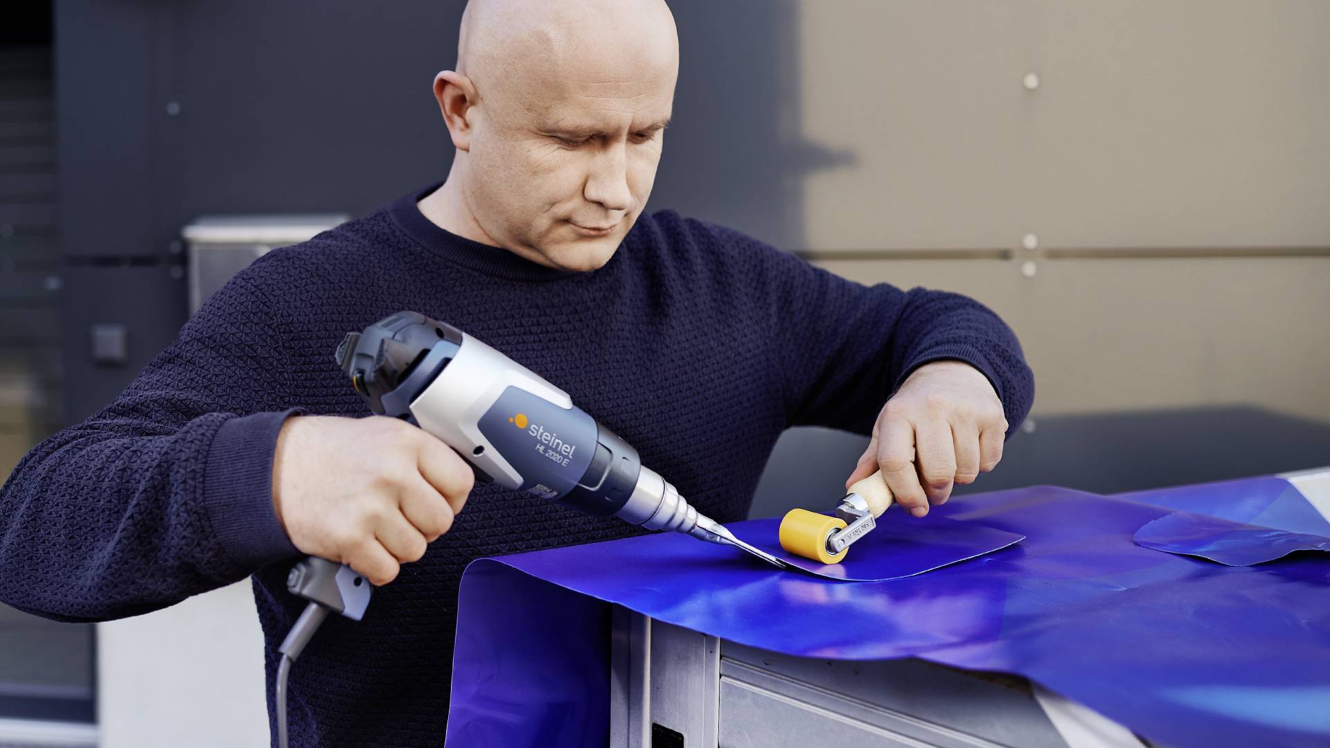 Ein Mann verwendet ein Elektrowerkzeug, um blaue Metallfolie auf einer Oberfläche zu bearbeiten. Er konzentriert sich auf seine Arbeit.