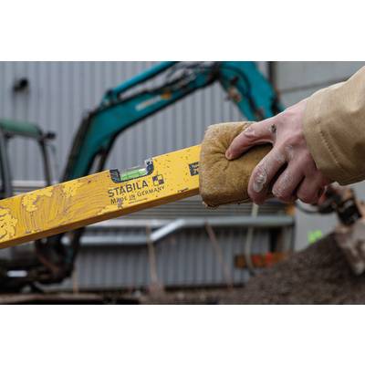 Eine Hand hält eine gelbe Wasserwaage ('Made in Germany') auf einer Baustelle. Im Hintergrund ist ein Bagger zu sehen.
