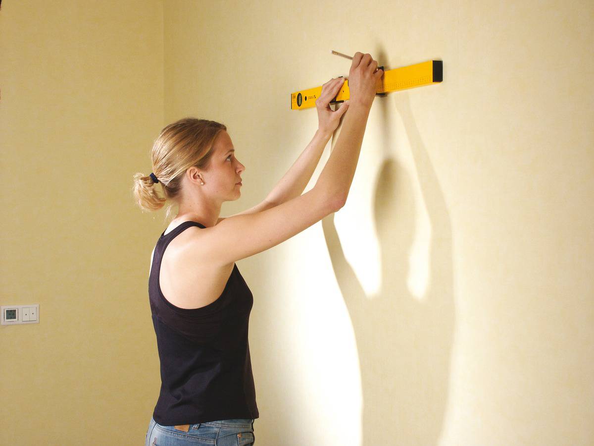 Eine Frau mit blondem Haar überprüft mit einer Wasserwaage, ob ein Punkt an einer Wand gerade ist.