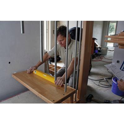 Ein Mann verwendet eine Wasserwaage, um ein Holzregal in einem Rohbau präzise auszurichten. Bauwerkzeuge und Materialien sind um ihn herum verteilt.
