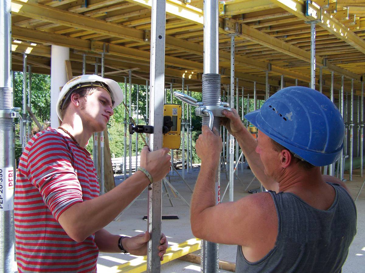 Zwei Bauarbeiter prüfen und justieren Gerüstteile auf einer Baustelle. Sie tragen Schutzhelme und arbeiten konzentriert.
